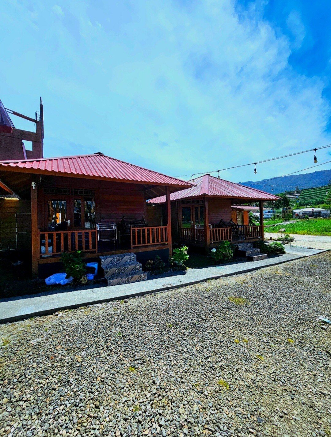 Two wooden cottages feature covered porches with railing, surrounded by gravel pathways. The roofs are red, complementing the natural landscape and greenery. A distant view of mountains can be seen under a clear blue sky, enhancing the tranquil setting.