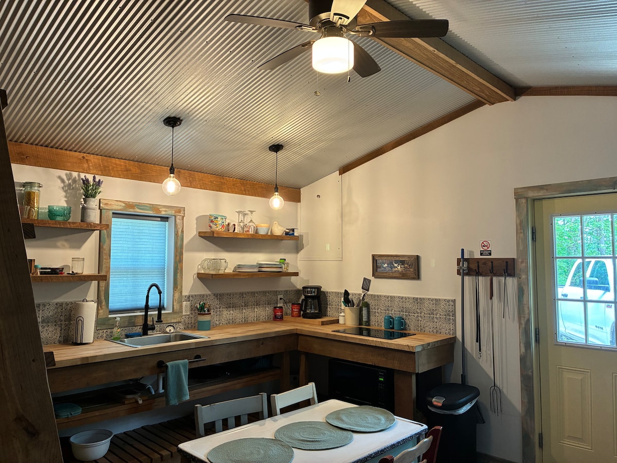 A functional kitchen area is equipped with wooden countertops and open shelving displaying dishware and cooking essentials. Pendant lights hang from the ceiling, and natural light enters through a window above the sink. A dining table with place settings is positioned nearby.