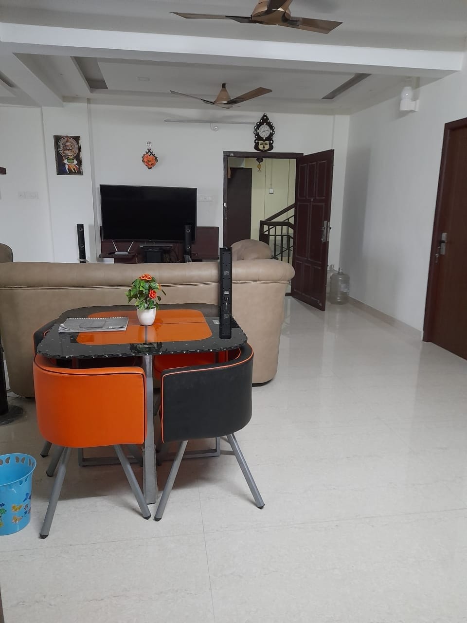 A combined living and dining area features a small table with orange and black chairs, accompanied by a decorative plant in the center. A large television is positioned against the wall, while a comfortable sofa provides seating. Bright tile flooring enhances the spacious atmosphere.
