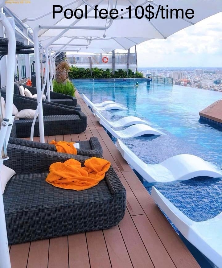 An infinity pool is featured on the rooftop, surrounded by stylish lounge chairs. Sunshades offer relief from sunlight while plush seating is accompanied by vibrant orange towels. The expansive view of the city skyline is visible in the background, creating a serene atmosphere.