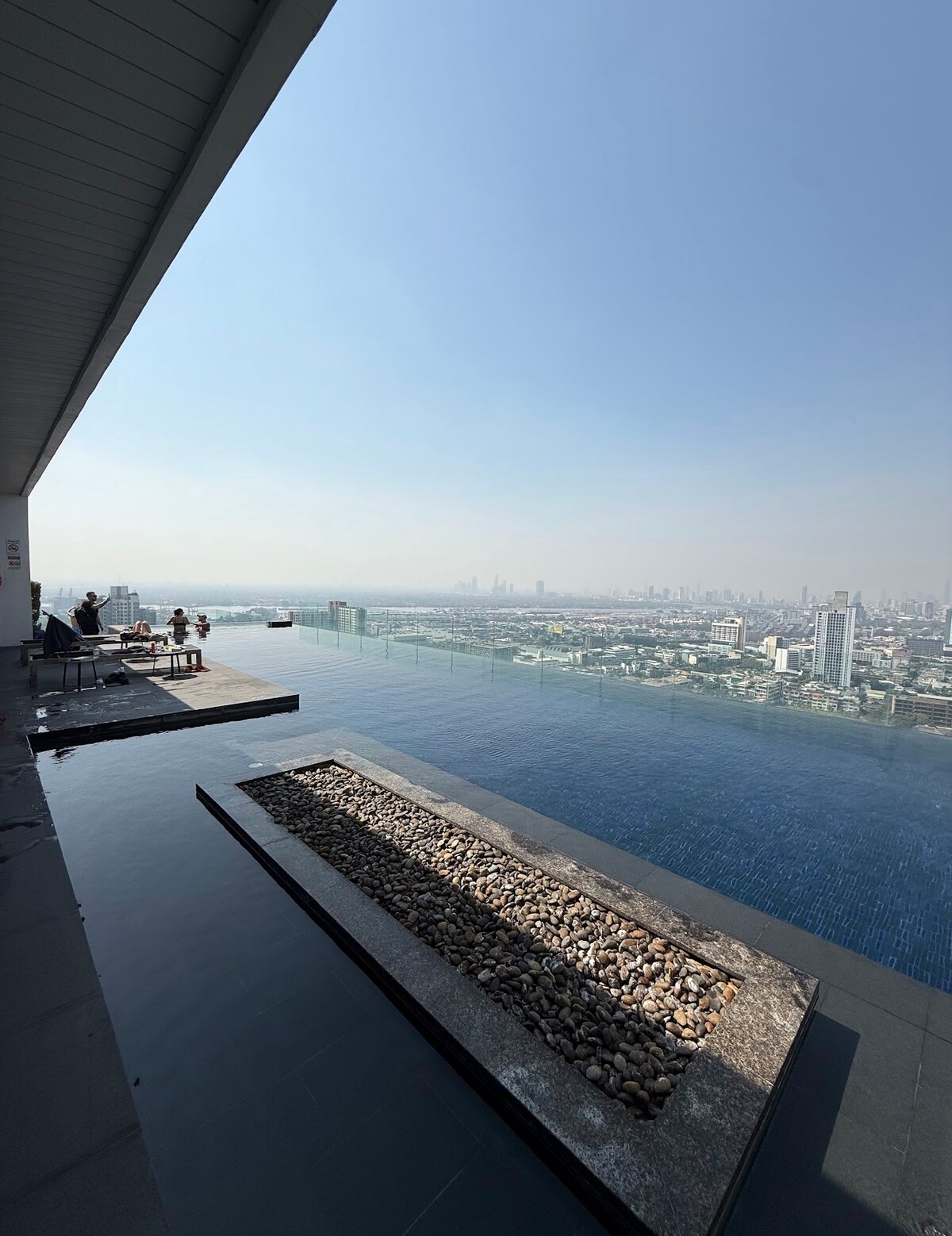 An infinity pool is positioned on the rooftop, providing uninterrupted views of the city skyline. The tranquil water reflects the clear sky, while a stone pathway runs alongside, leading to a lounge area with chairs. The setting offers a peaceful atmosphere for relaxation.