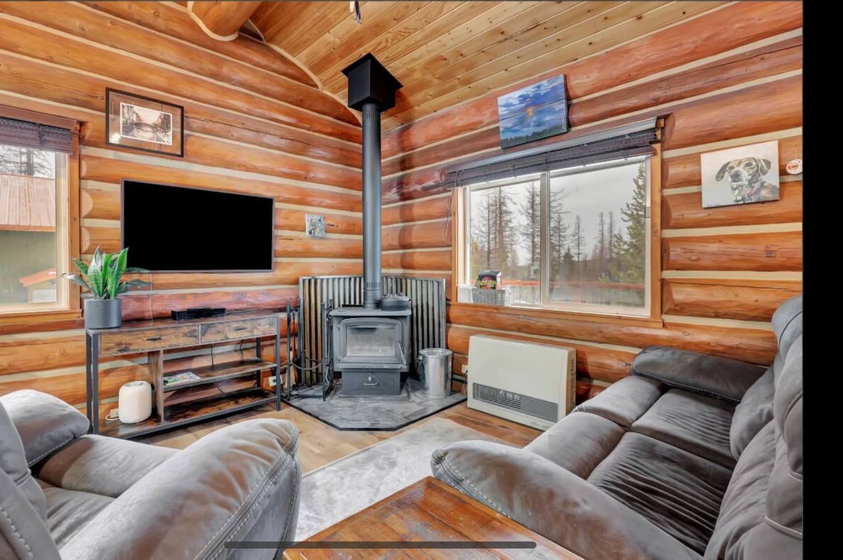 A cozy living area is situated within a log cabin, featuring a plush seating arrangement and a wood stove. A large window offers views of the surrounding trees, while a modern television mounted on the wall complements the warm wooden interior.