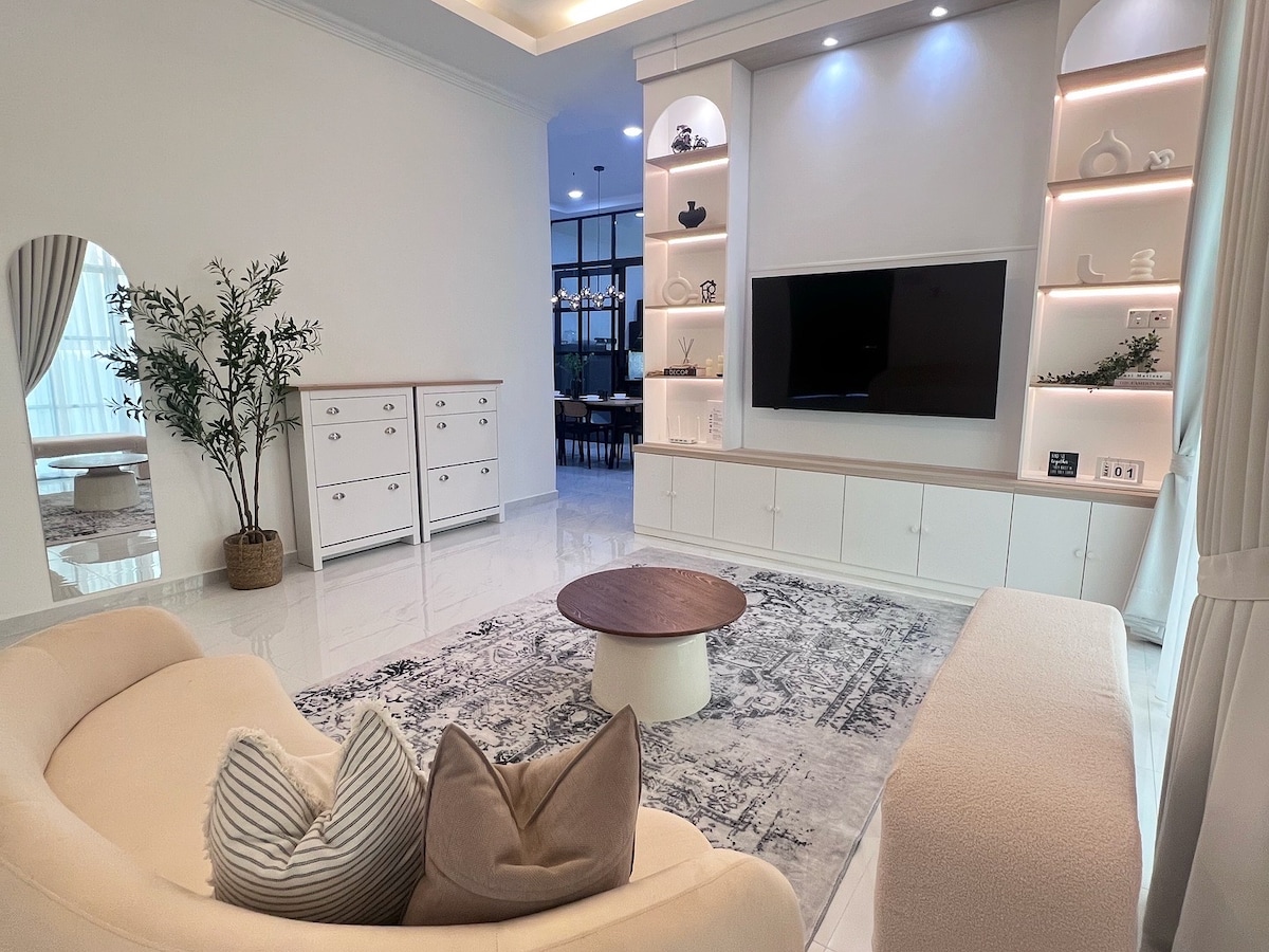 A spacious living area is featured, highlighted by a cream-colored sofa and a circular coffee table atop a soft area rug. The wall-mounted TV is surrounded by built-in shelves that display decorative items, while large windows invite natural light into the bright room.