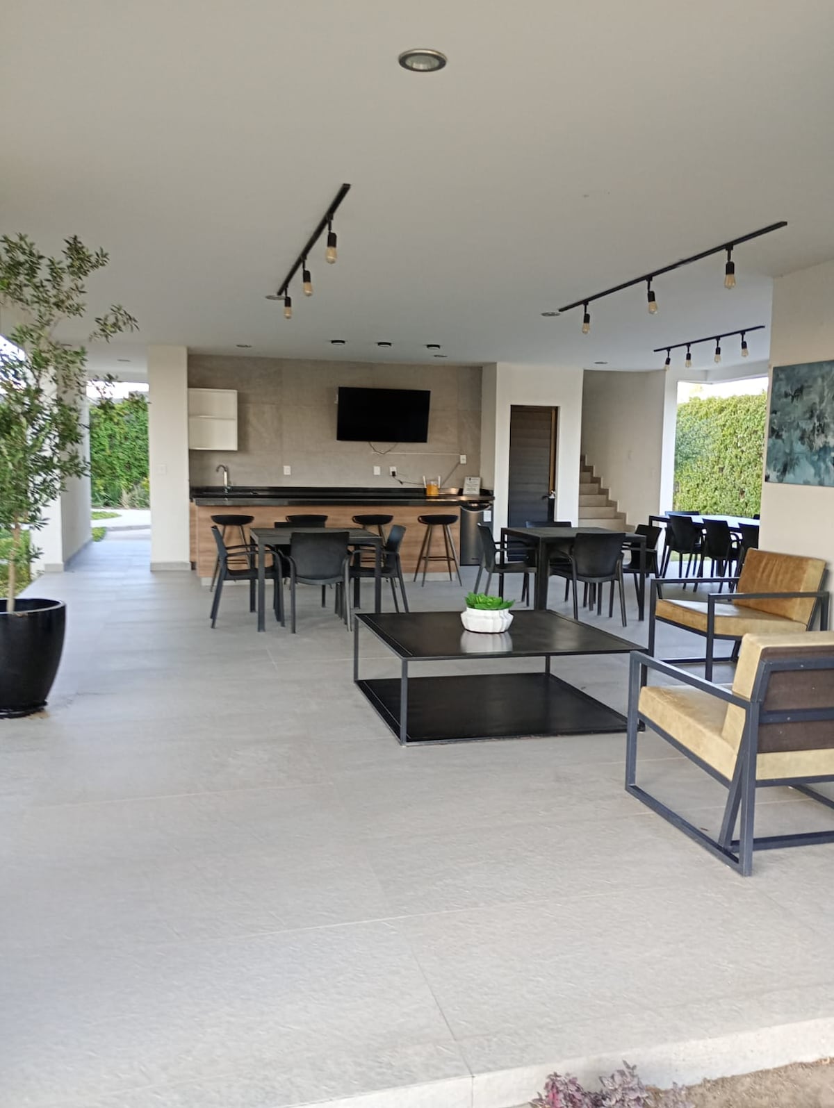 A spacious communal area is presented with a seating arrangement featuring black metal chairs and a low table, complemented by wooden armchairs. An outdoor kitchen is visible, along with a mounted television. Natural light fills the space through large windows, enhancing the modern, minimalistic design.