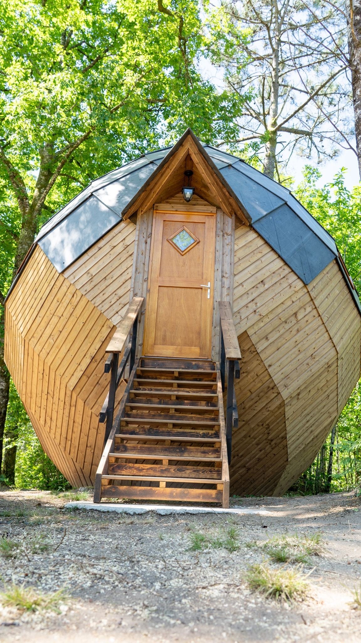 A unique Zome structure is presented, featuring an angular wooden exterior and a distinctive geometric design. Stairs lead up to a charming wooden door adorned with a diamond-shaped window, surrounded by lush trees, creating a serene natural environment.