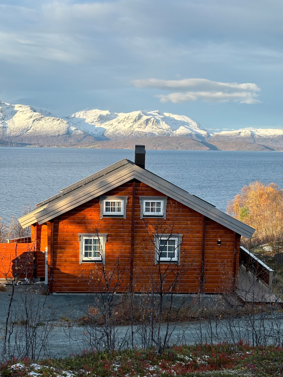 A rustic wooden cabin is nestled against a backdrop of snow-capped mountains and a tranquil lake. Large windows reflect the surrounding nature, while the vibrant red exterior creates a warm contrast with the cool landscape. The setting invokes a sense of peace and solitude.