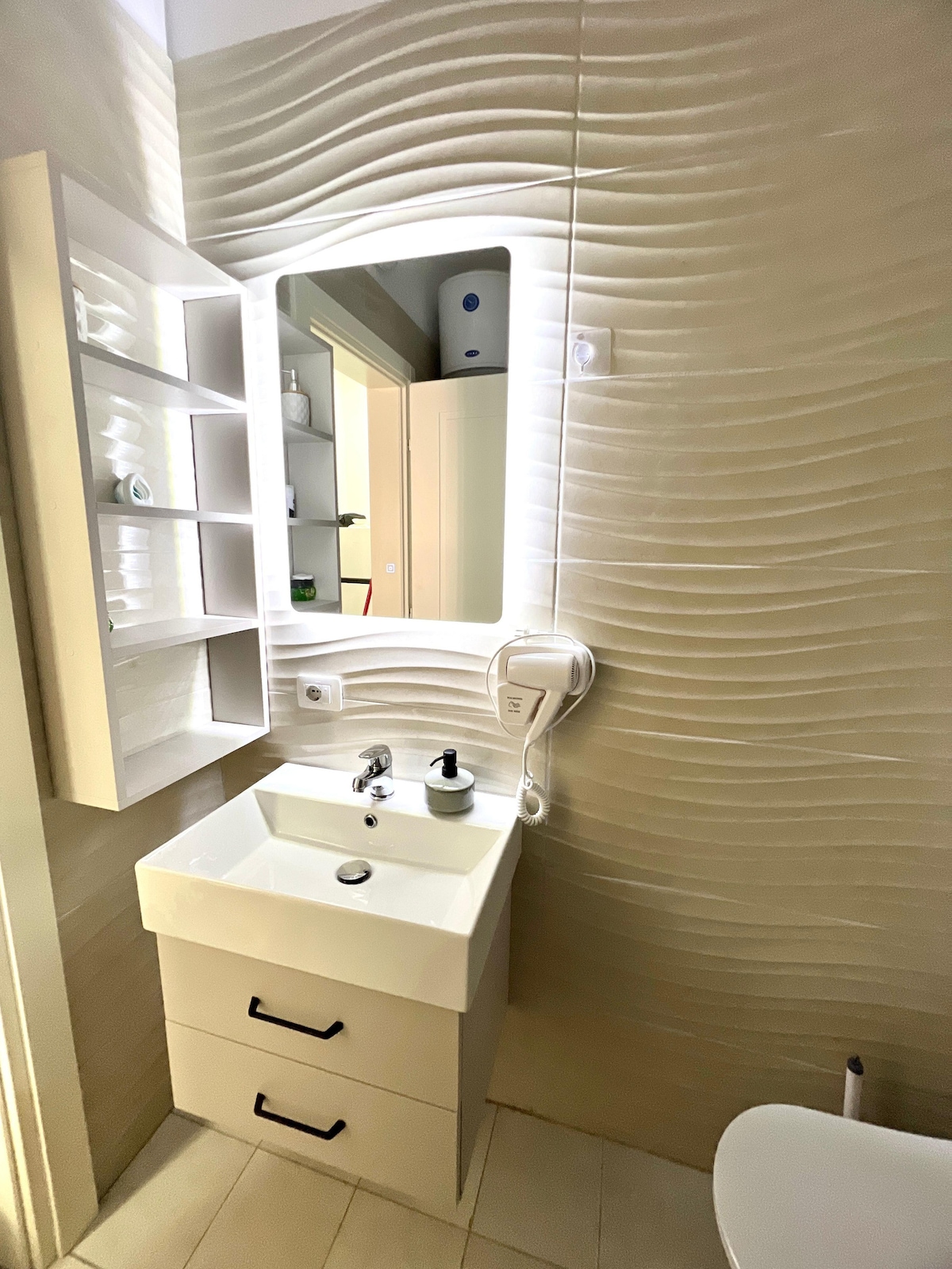A modern bathroom is displayed, featuring a sleek sink with a minimalist design. A large mirror with soft lighting enhances the space, while a wall-mounted shelf in a neutral tone showcases neatly arranged toiletries. Textured wall panels add an element of depth to the decor.
