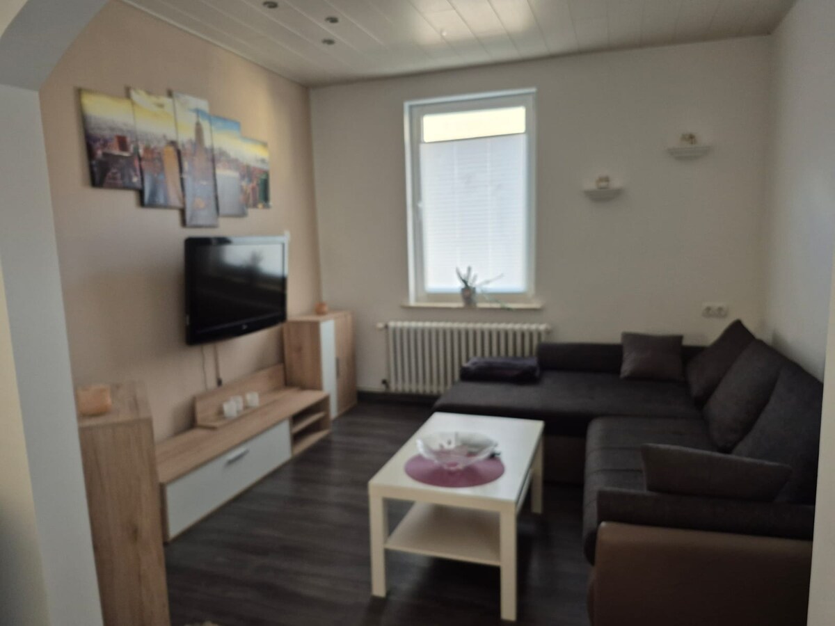 A comfortable living area is presented with a large sectional sofa and a coffee table. A mounted television is positioned on a light wood entertainment unit. The space is brightened by a window with light filtering through and decorative shelves featuring simple ornaments.