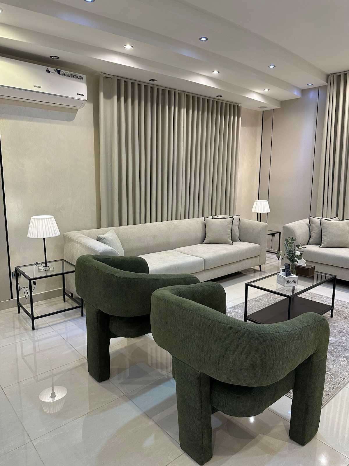 A modern living area is presented with a comfortable sofa and two uniquely shaped armchairs. A small coffee table is centered on a soft rug, complemented by decorative lamps. Light is softly diffused from recessed lighting above and natural light coming from outside.