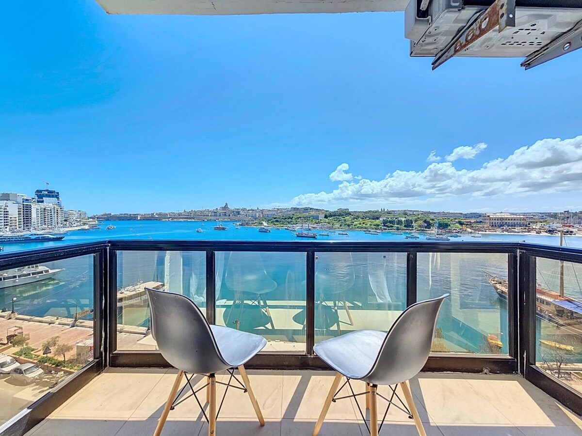 A balcony with two modern chairs overlooks the shimmering Mediterranean Sea, providing a clear view of Valletta's iconic skyline. The bright blue sky is dotted with a few clouds, enhancing the serene atmosphere of this coastal location.