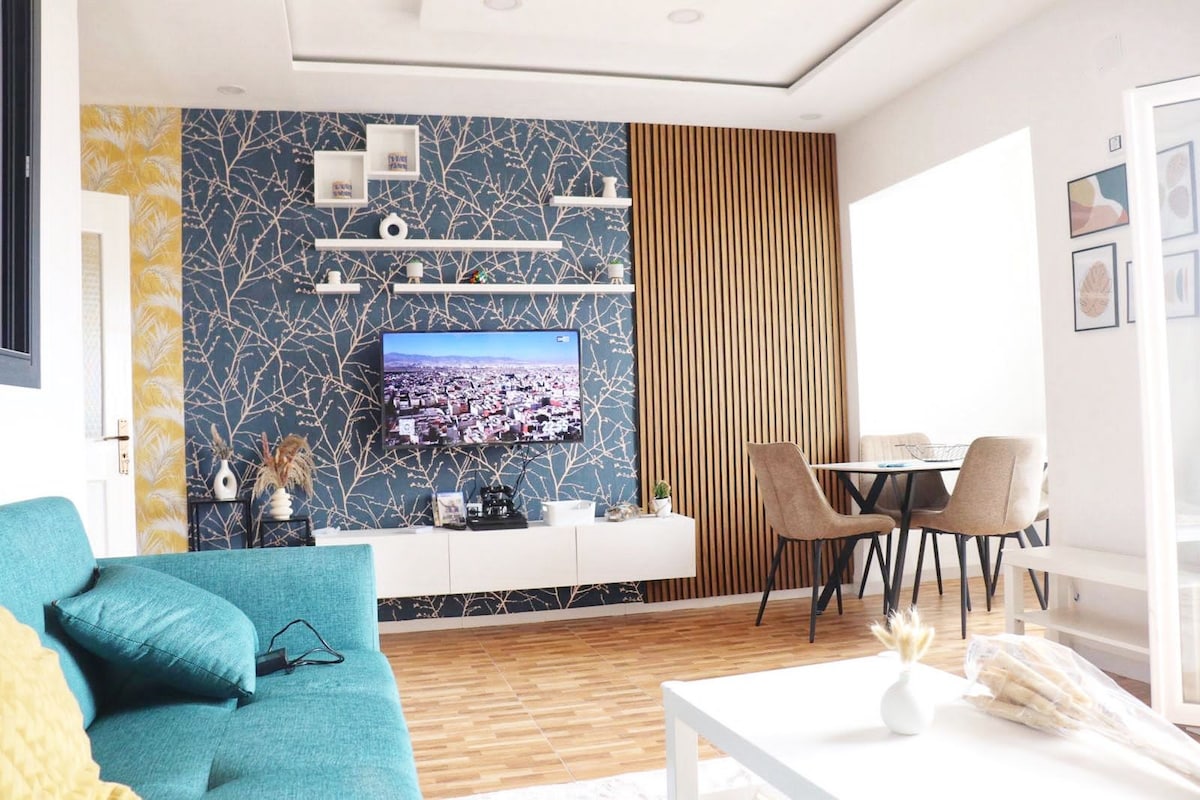 A modern living area is showcased with a bright blue accent wall adorned with a branch pattern. A flat-screen TV is mounted above a sleek white media unit. A light wood dining table is set for four, and a comfortable teal sofa is positioned to create a welcoming space.