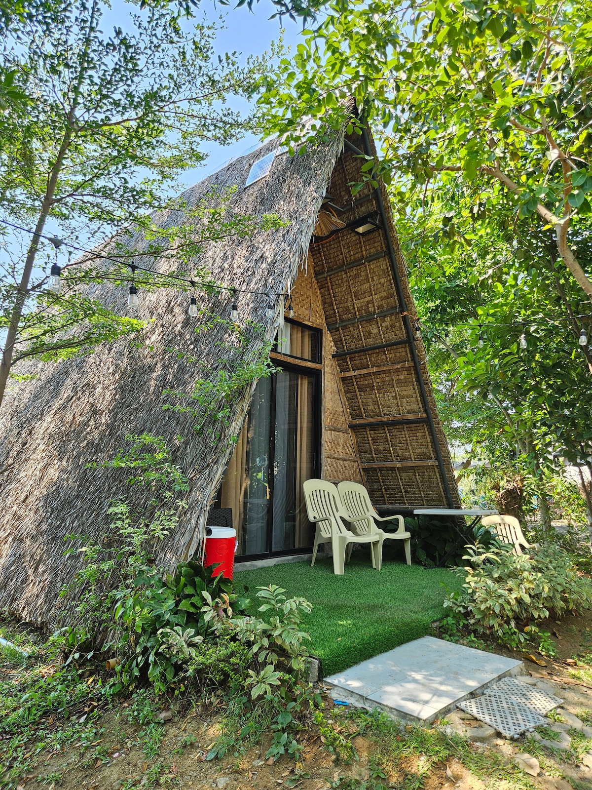 A unique cabin with a distinctive thatched roof sits among lush greenery. Two light-colored chairs are positioned on a small patio, accompanied by a table. A cooler is placed nearby, providing convenience for outdoor activities. The overall setting evokes a calm and serene environment.