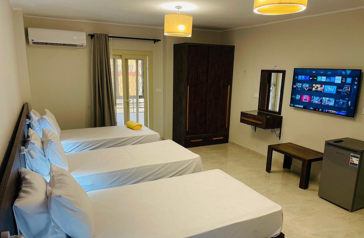 A spacious room is presented with three beds dressed in white linens. A flat-screen television is mounted on the wall opposite, while a mini fridge and a wooden table are positioned nearby. Natural light enters through a set of double doors, which lead to a balcony.