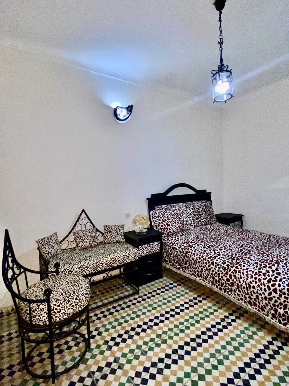 A cozy bedroom features a spacious bed dressed in a leopard-print blanket. A patterned tiled floor adds visual interest. A small seating area, including a bench and a chair with similar upholstery, is positioned near the bed, illuminated by unique pendant lighting.