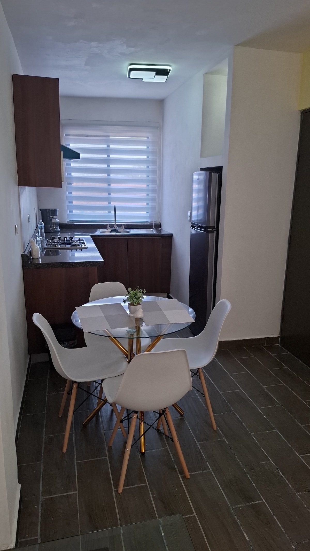A compact kitchen area is equipped with modern appliances and wooden cabinetry. A round dining table with four white chairs is positioned near a window, allowing natural light to brighten the space. The flooring features stylish dark tiles, contributing to a contemporary look.