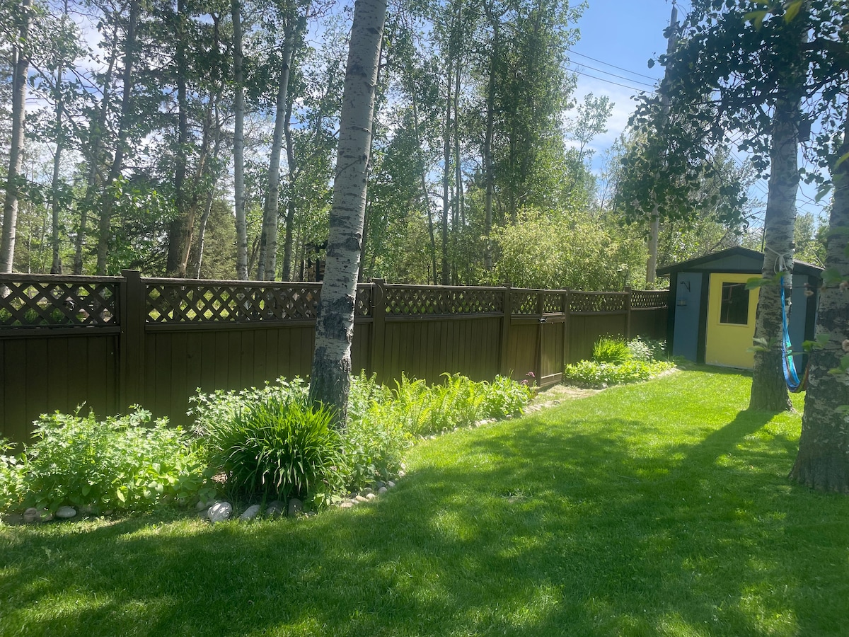 A green backyard is surrounded by tall trees and a wooden fence, providing privacy. Lush garden beds filled with various plants line the pathway, which leads to a small, yellow shed. Sunlight filters through the trees, creating serene dappled lighting on the grass.