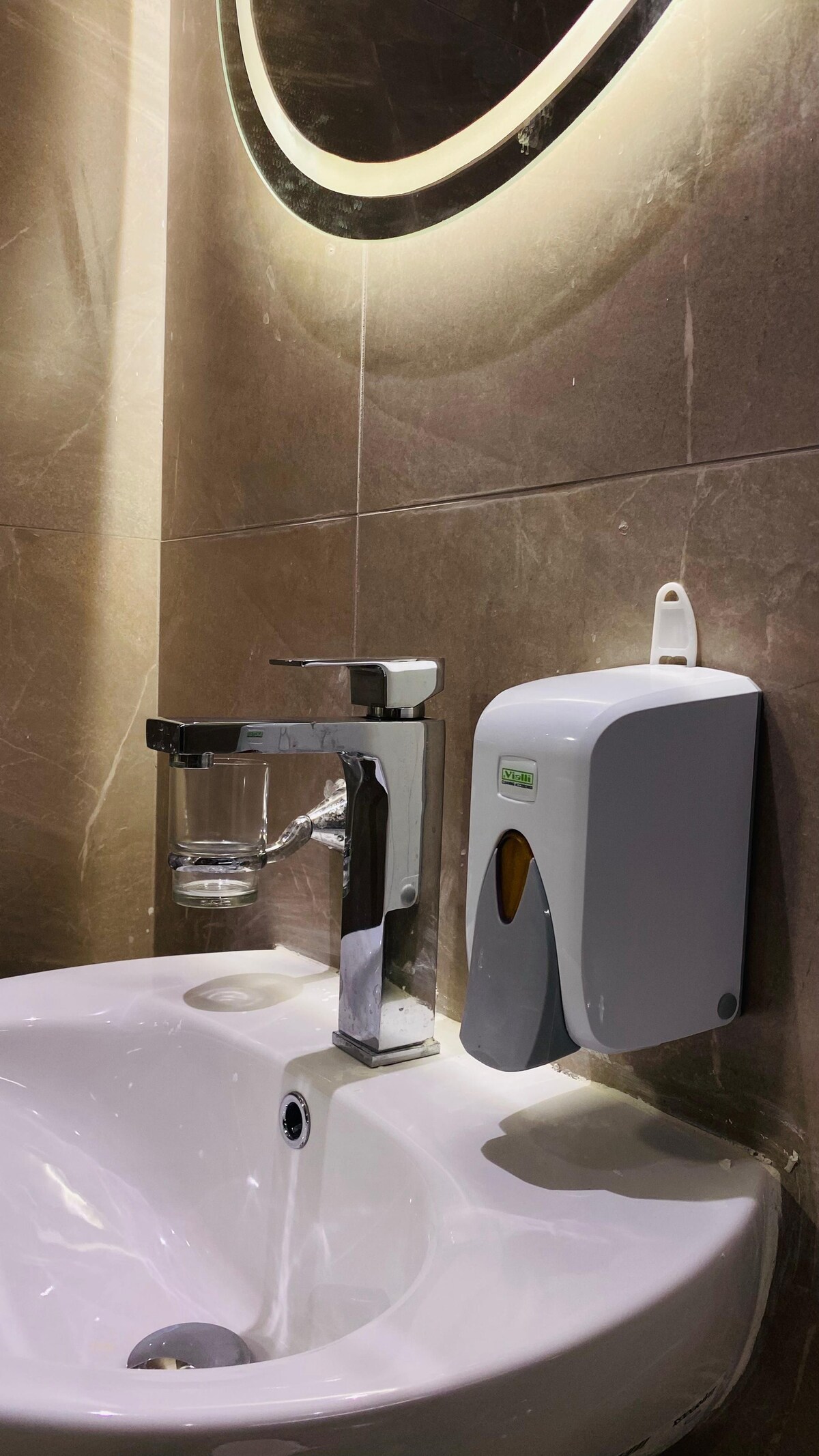 A neatly designed bathroom sink area is highlighted, featuring a modern faucet with a glass tumbler. A wall-mounted soap dispenser is positioned beside the sink, which is set against elegant, textured tile walls illuminated by soft lighting.