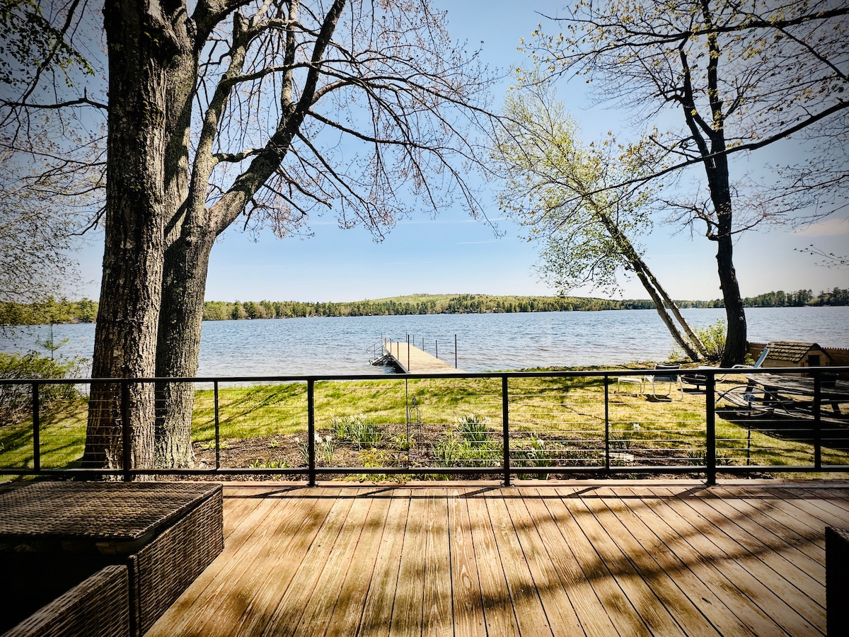 The image captures a serene view from a deck, showcasing a calm lake with a wooden dock extending into the water. Lush green grass surrounds the area, with trees framing the scene, providing a peaceful outdoor setting.