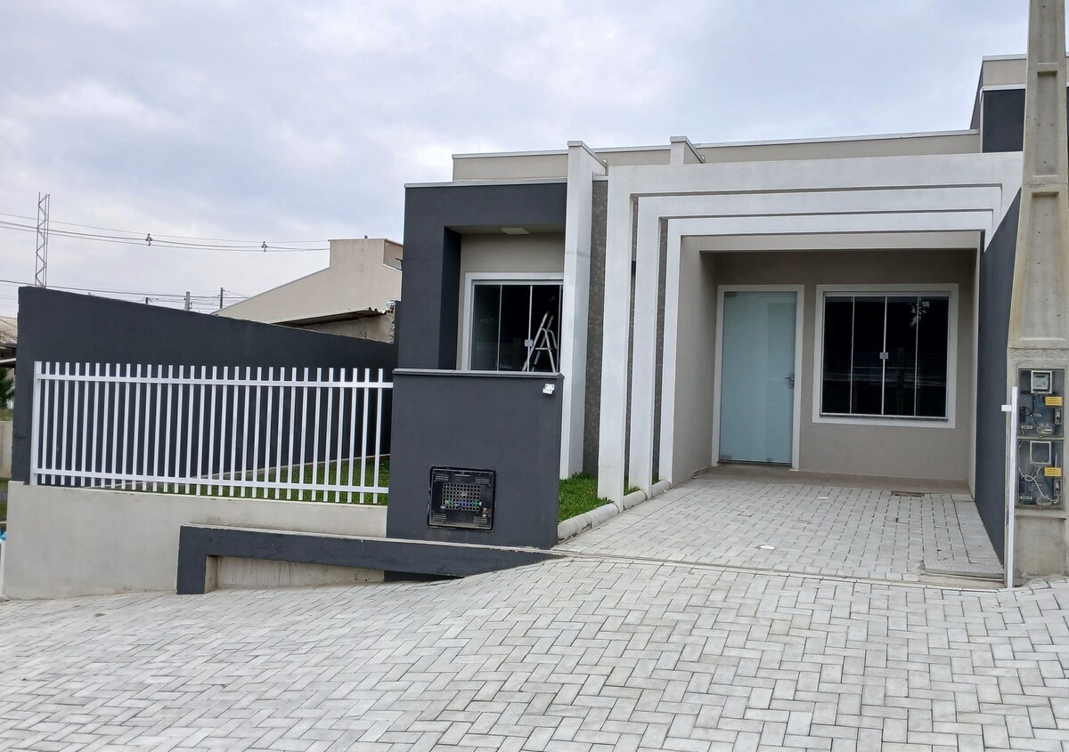 A modern one-story house is shown with a clean exterior design. A spacious driveway leads up to the entryway, featuring large windows that allow natural light to fill the interior. A fenced area is visible, providing a sense of privacy.
