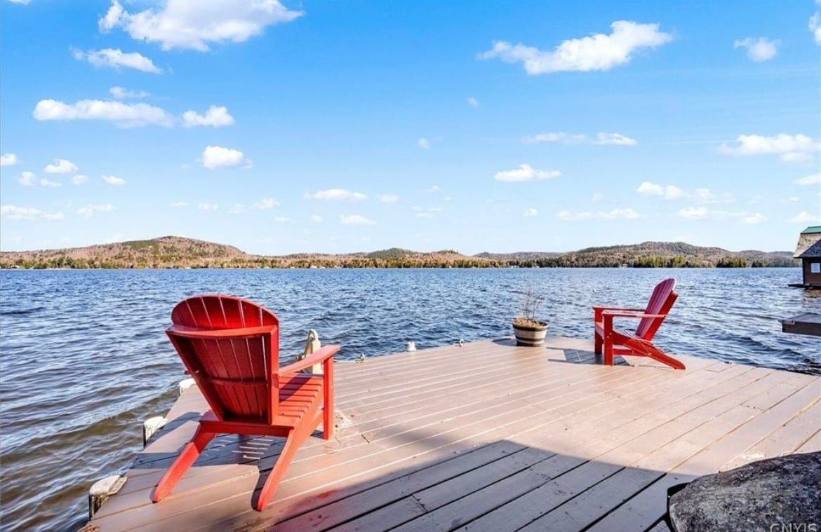 Two red Adirondack chairs are positioned on a wooden dock overlooking a calm lake. The serene water is surrounded by rolling hills under a clear blue sky, with scattered clouds enhancing the outdoor scene.