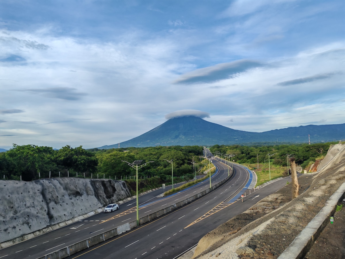 A serene view of a highway, bordered by lush greenery, leads towards a majestic volcano shrouded in clouds. The roadway is smooth and appears well-maintained, while the mountainous landscape rises in the background, creating a tranquil scene in a natural setting.