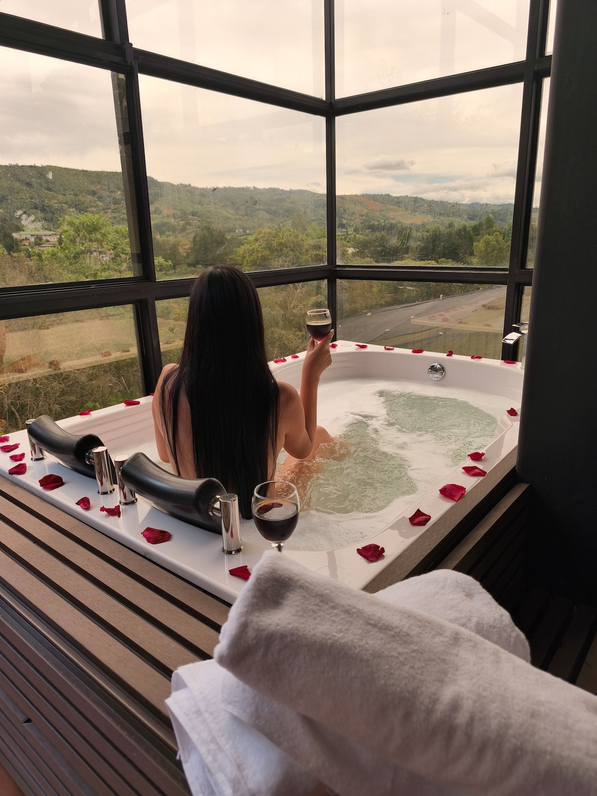 A person is seen enjoying a jacuzzi surrounded by glass windows, offering a view of the mountains and landscape. Rose petals are scattered around the tub, enhancing the serene ambiance. A towel is neatly folded nearby, providing a touch of comfort and convenience.