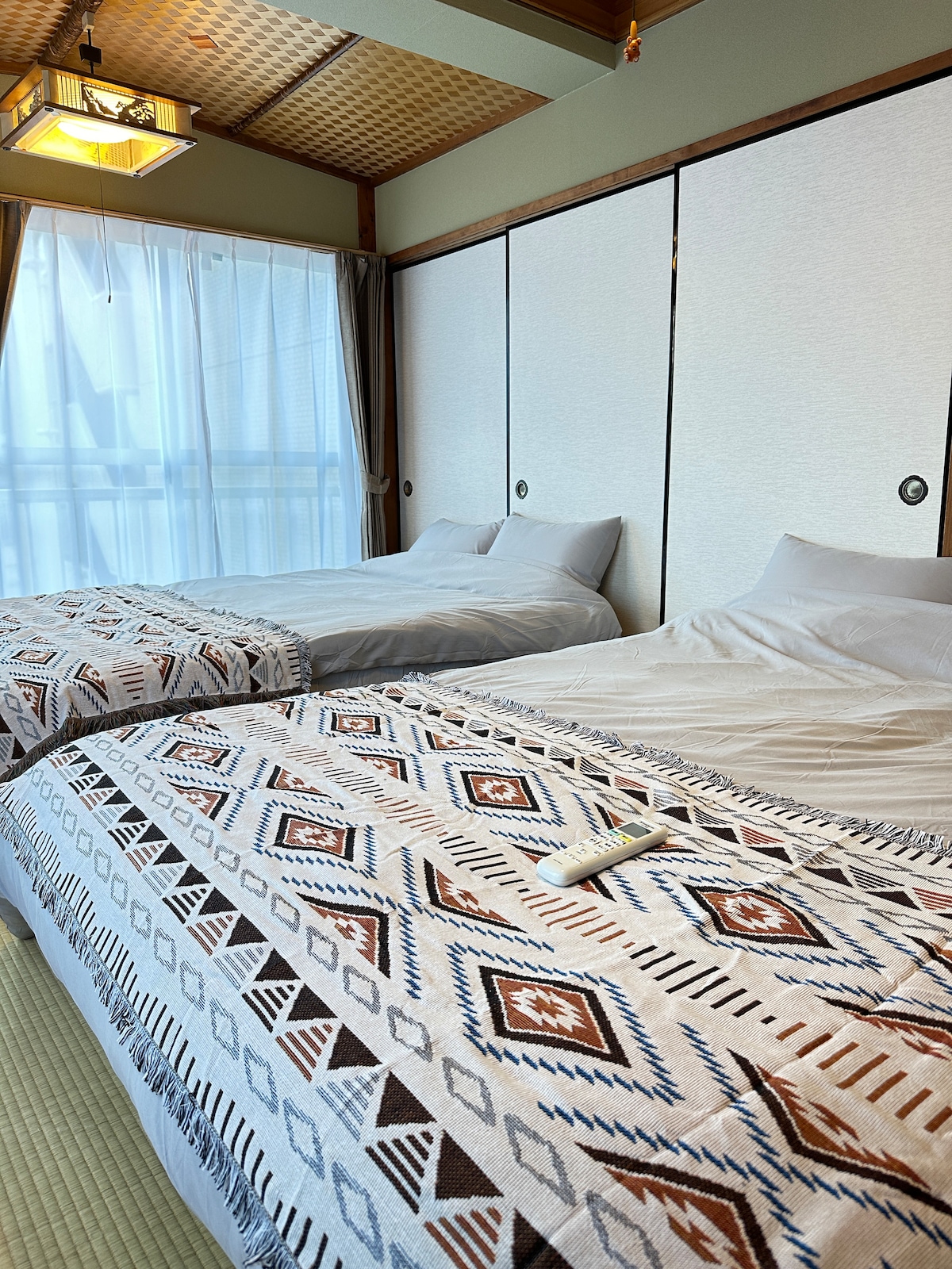 The room features two spacious beds dressed in light-colored linens, complemented by a patterned blanket that adds texture. Natural light filters through sheer curtains, illuminating the minimalist decor with sliding panel walls, creating a calming atmosphere.