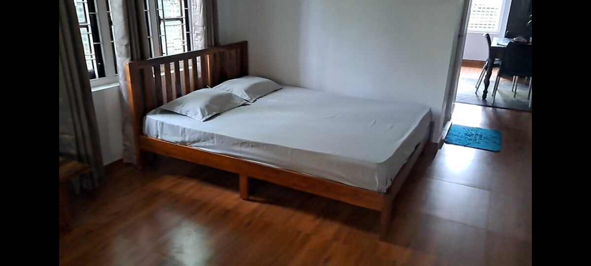 The room features a spacious wooden bed frame with a neutral-colored mattress. Natural light filters through the nearby windows, illuminating the polished wood flooring. In the background, a dining area is visible, adding depth to the setting.