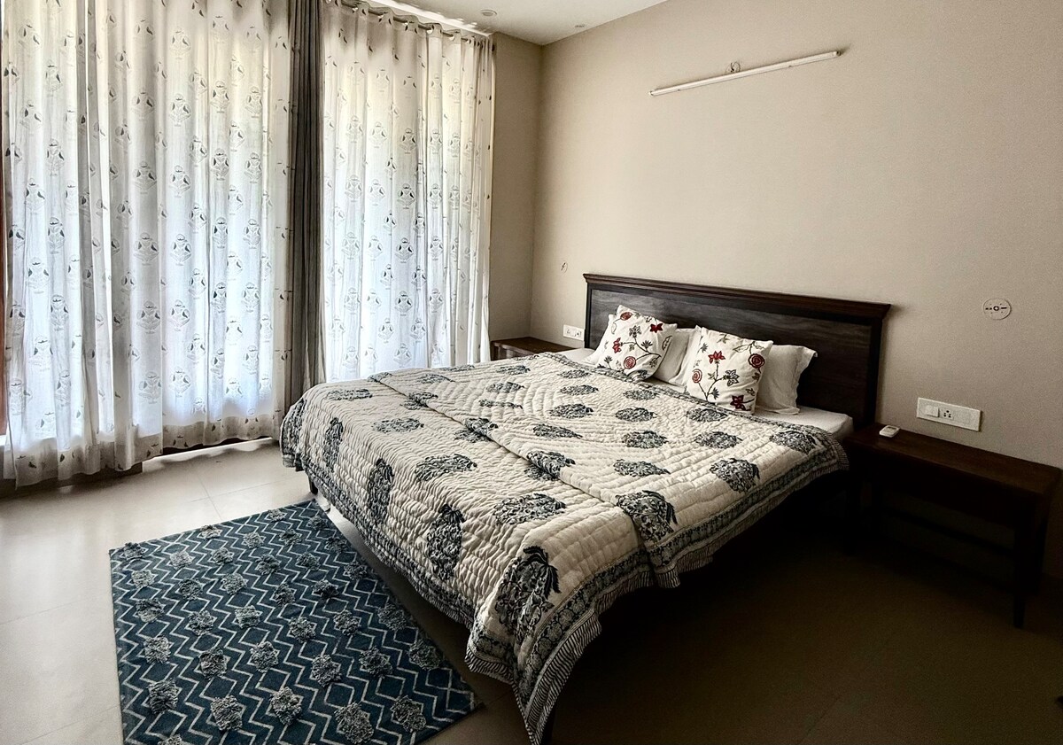 A spacious bedroom features a large bed with decorative pillows. Natural light filters through sheer curtains, creating a soft glow. A patterned rug adds texture to the floor, while a simple wooden nightstand complements the minimalist design.