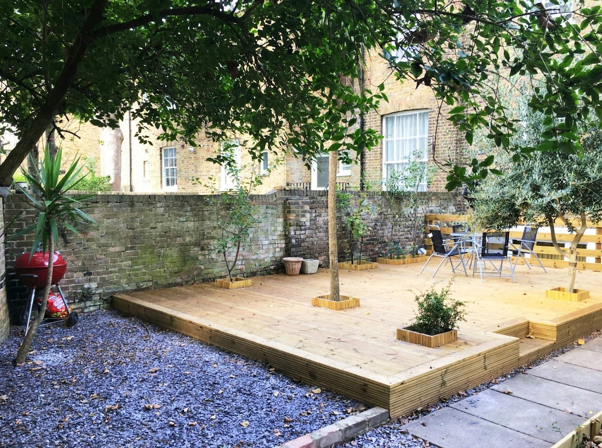 An outdoor space features a wooden deck surrounded by greenery and stone pathways. A circular dining table is set with four chairs, and a grill is positioned nearby. The background displays a brick wall and several trees, enhancing the natural setting.