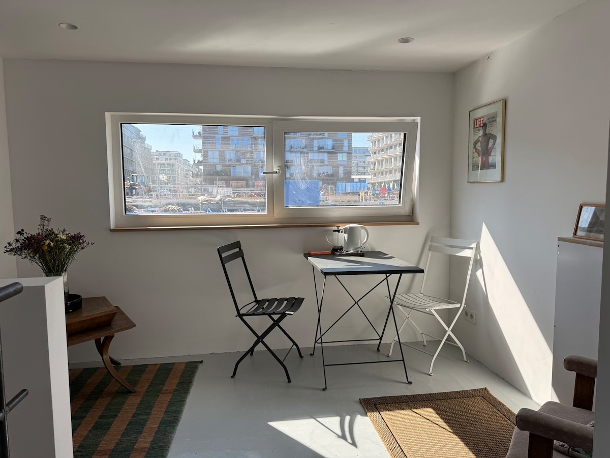 A bright living area features two chairs and a small folding table by large windows allowing natural light to fill the space. A kettle sits on the table, with simple decor enhancing the minimalist aesthetic. The view includes nearby buildings and outdoor scenery.