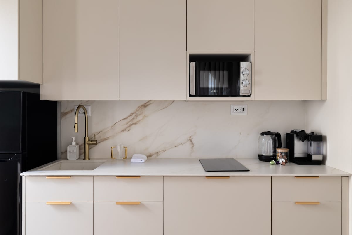 A modern kitchen features sleek cabinetry with gold handles, a white countertop, and a marble backsplash. A microwave is mounted above the counter, accompanied by a kettle and coffee maker. A black refrigerator stands to the left, providing a clean and organized cooking space.
