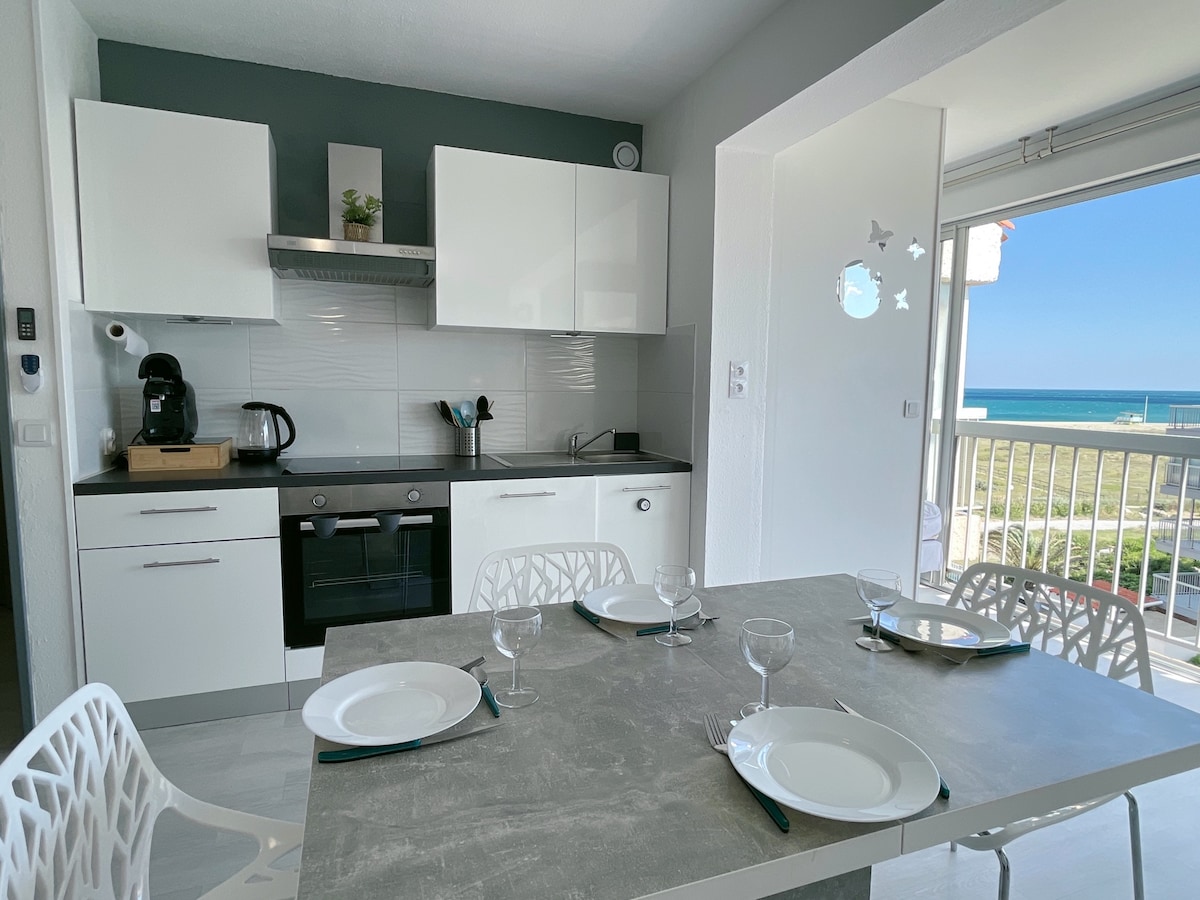 A modern kitchen area features sleek white cabinetry and a contrasting dark countertop. A dining table is set for four with plates and glasses. A sliding glass door invites natural light and provides a view of the nearby beach and sea.