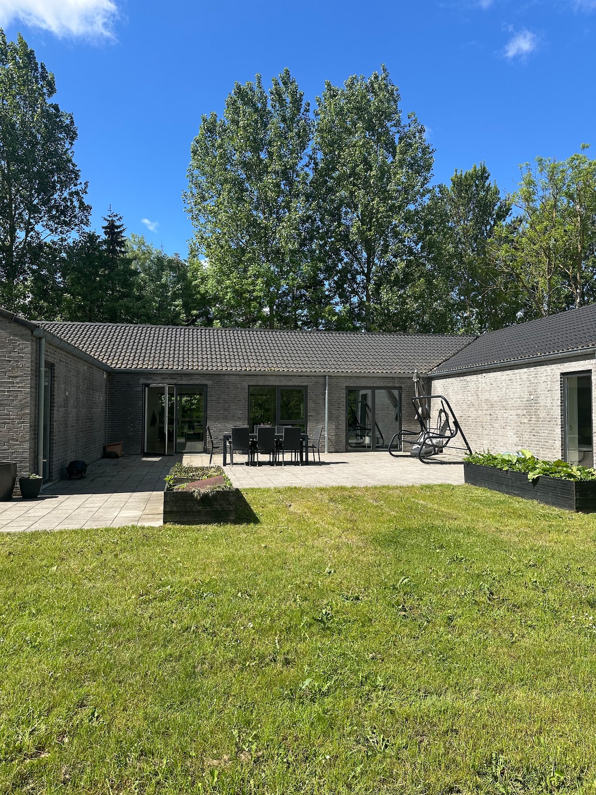 A spacious outdoor area is viewed, featuring a well-maintained lawn and paved patio. Several black outdoor chairs surround a large table, providing an ideal setting for gatherings. Bright blue skies and tall trees create a serene backdrop for this inviting space.