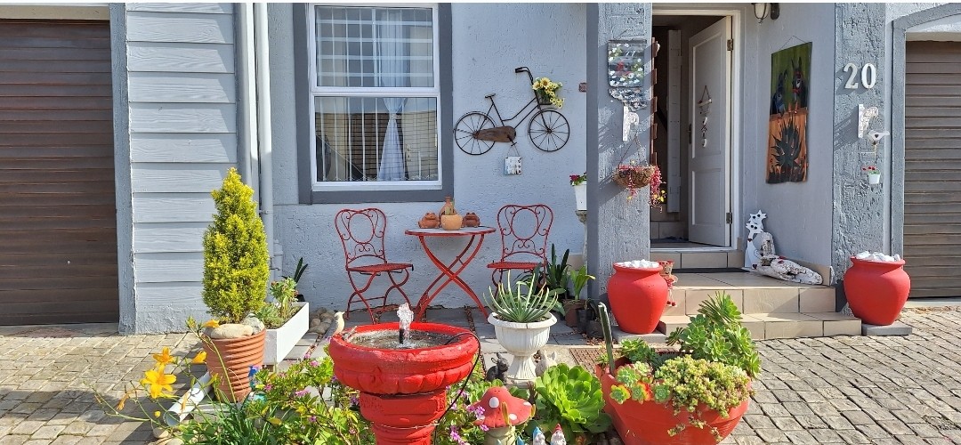 The entrance of the townhouse features a small outdoor seating area with two red chairs and a round table, surrounded by colorful pots of plants and flowers. A bicycle art piece is mounted on the wall, enhancing the welcoming ambiance.