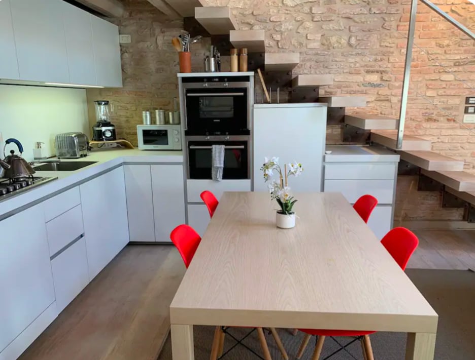 A modern kitchen area features a wooden table surrounded by six red chairs. A fully equipped kitchen with sleek cabinetry includes a built-in oven and microwave, alongside a kettle and coffee maker. Stone walls add texture, with wooden stairs leading to an upper level.