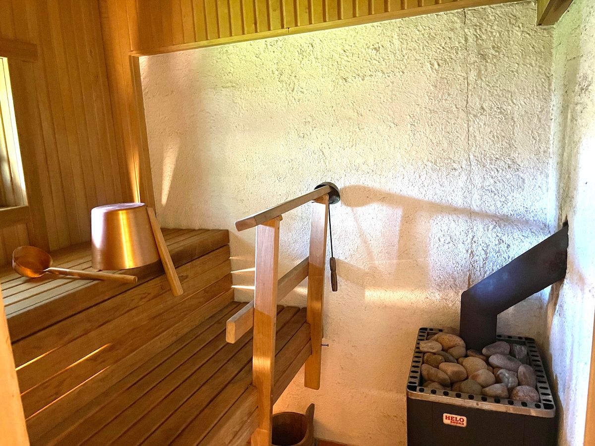 The interior of the sauna features wooden benches arranged for seating, with a metal bucket and ladle placed nearby. A stove filled with stones is positioned in the corner, providing heating for the space. Natural light filters through a small window.