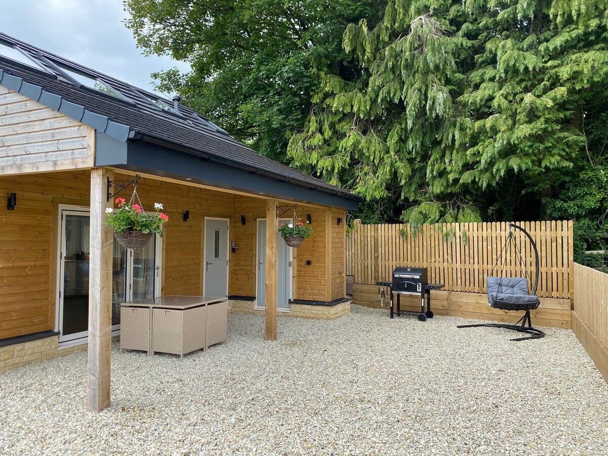 An outdoor space features a light gravel surface surrounded by wooden fencing. Two hanging flower baskets add color near a seating area with woven furniture. A charcoal BBQ grill is positioned nearby, alongside a freestanding swinging chair, inviting relaxation in the fresh air.