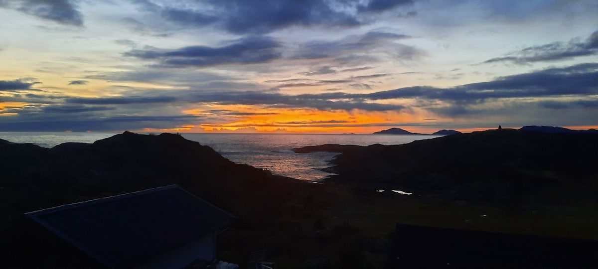 A scenic view of the ocean at sunset is captured, featuring vibrant orange and purple hues across the sky. Silhouettes of distant islands and rocky coastline are visible against the water's reflective surface, creating a calm and serene landscape.