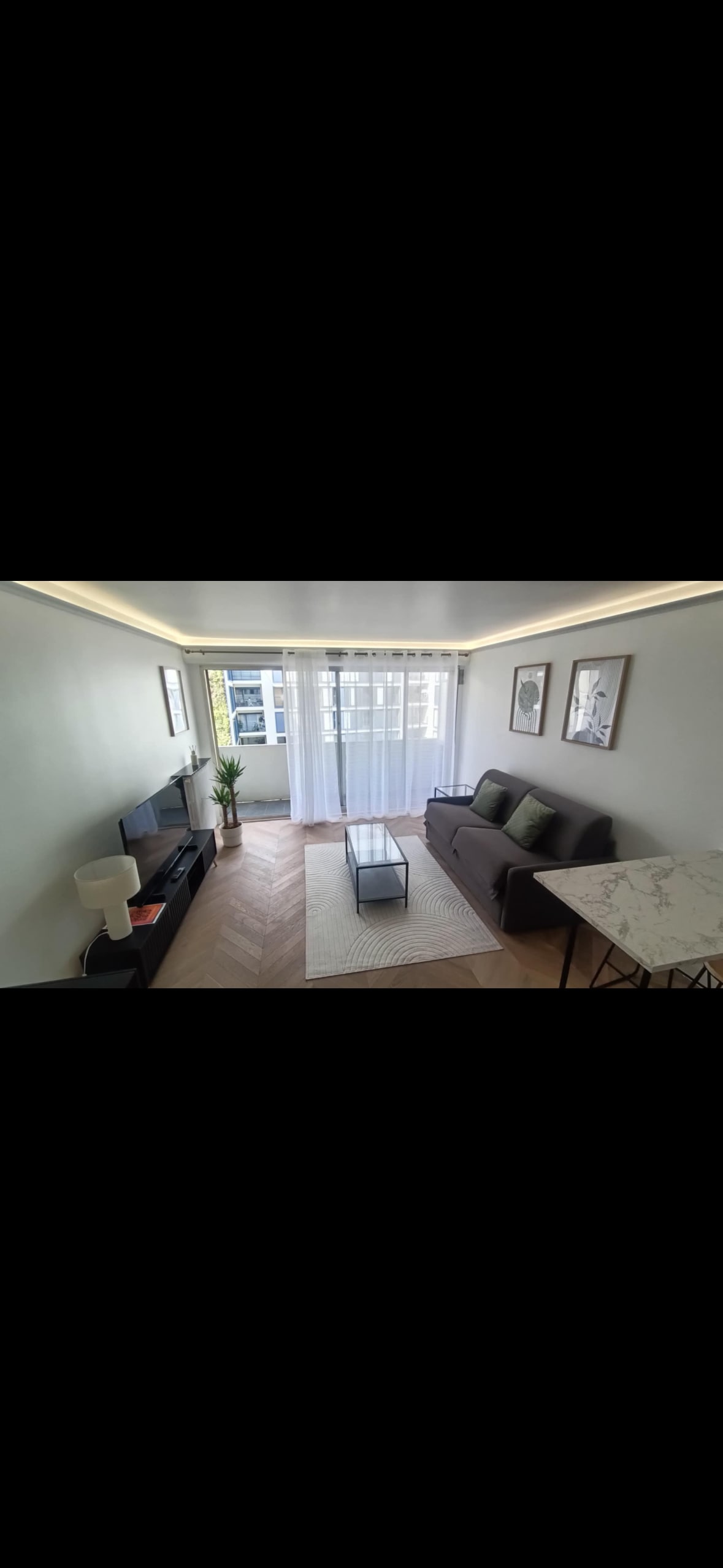 A bright living space features a comfortable dark sofa with green accent pillows. A coffee table sits on a light area rug, complemented by a console table with a television. Large windows allow natural light to fill the room, while framed artwork adorns the walls.