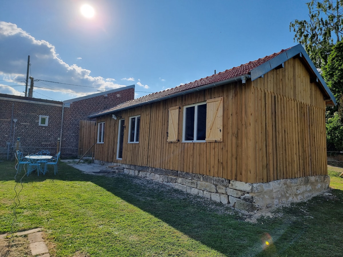 A wooden house with a rustic exterior is showcased under bright sunlight. The structure features multiple windows with wooden shutters, and a door on the front. A grassy area surrounds the house, complemented by a blue outdoor table and chairs nearby.