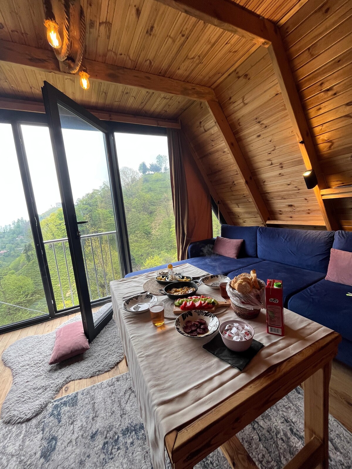 A wooden dining table is set with various dishes, including fruits and beverages. A spacious blue sofa provides seating, and large glass doors lead to a balcony, offering a view of green hills. Warm wooden paneling adorns the walls and ceiling.