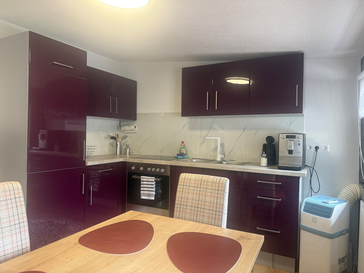 A modern kitchen is presented with dark purple cabinetry and a light countertop. Essential appliances, including an oven and coffee maker, are visible. A wooden dining table with patterned chairs occupies the space, creating a functional area for meals.