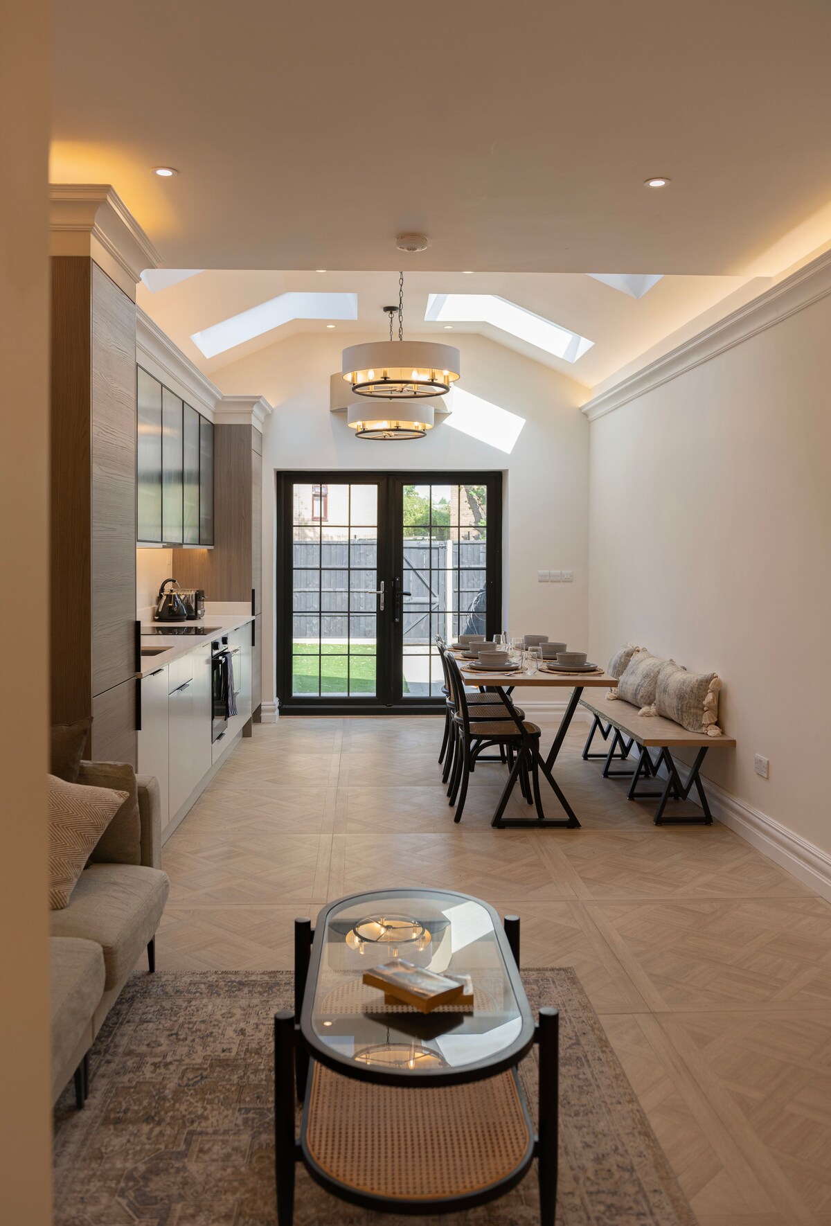 An airy open-plan dining and living area is highlighted by a combination of skylights and large windows. A contemporary dining table is set for several guests, while a cozy seating area includes a subtle rug and a stylish coffee table. The inviting garden can be seen through the glass doors.