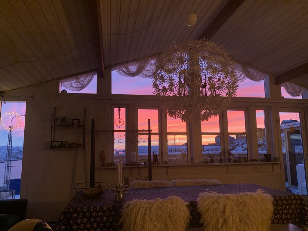 A dining area is featured, illuminated by a striking sunset view through large windows. A decorative chandelier hangs from the ceiling, while a table adorned with a patterned tablecloth is surrounded by cozy seating. Soft textures are present, adding warmth to the space.