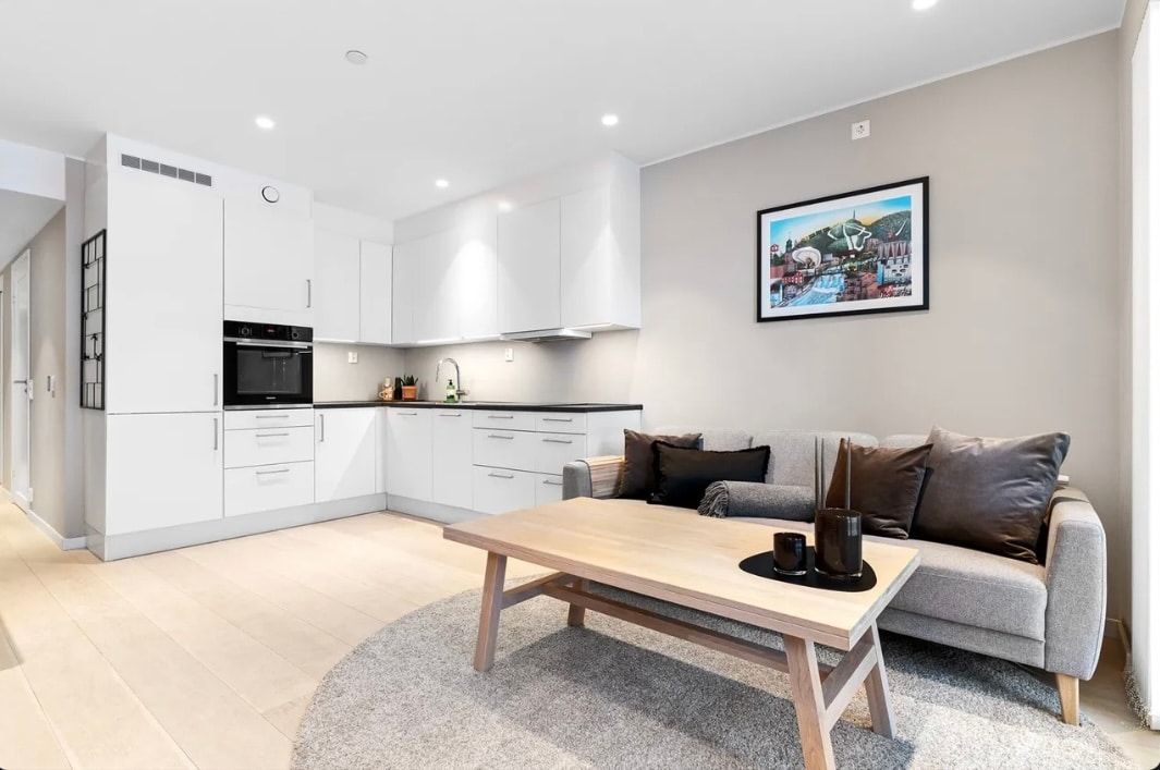 A modern kitchen area is complemented by sleek white cabinetry and built-in appliances. A cozy seating area features a light gray couch adorned with dark cushions, positioned next to a wooden coffee table. Soft lighting enhances the neutral color palette of the space.
