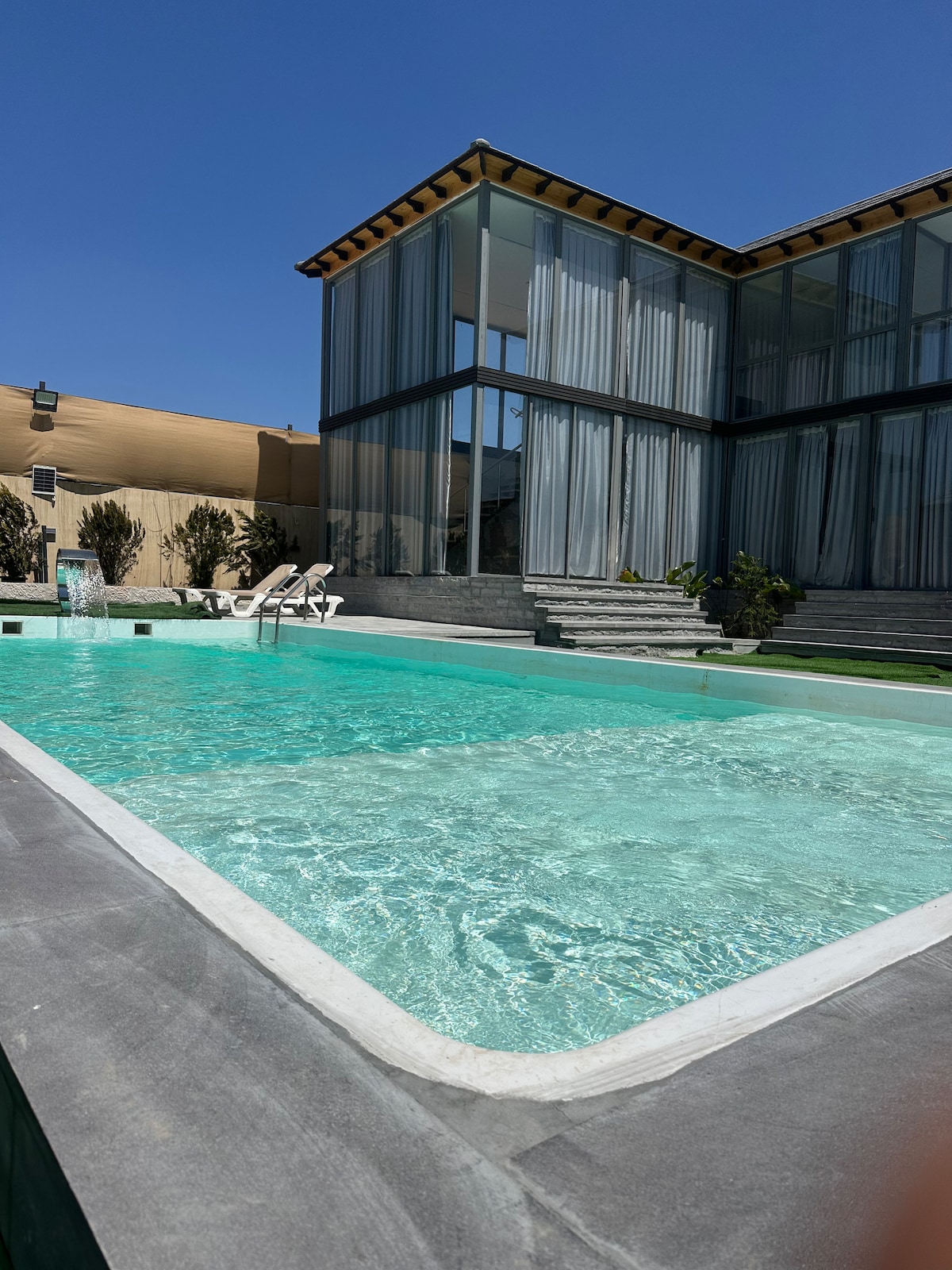 An outdoor pool is featured with clear turquoise water, surrounded by light-colored stone. The modern multi-story building, noted for its extensive glass windows, showcases a contemporary design. A few sun loungers are visible beside the pool, enhancing the inviting setting.