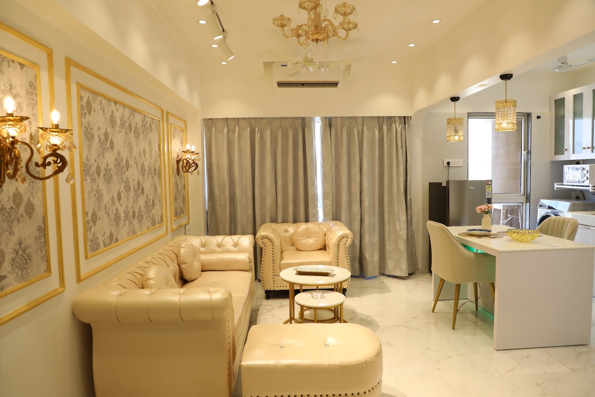 A stylish living area is showcased with elegant beige furnishings, including a plush sofa and armchairs. Soft lighting illuminates the space, accentuating the intricate wall detailing and large windows covered with light gray curtains. A dining table and kitchen area are visible in the background.