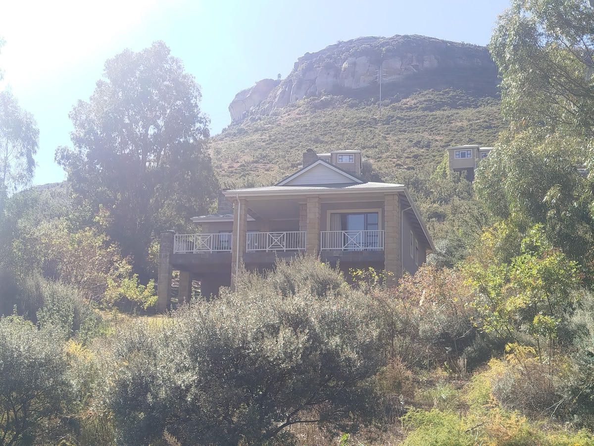A charming villa is set against a backdrop of mountains, surrounded by lush greenery. The spacious front porch is supported by sturdy pillars, and the facade features a combination of stone and wood. Natural light filters through the trees, enhancing the serene atmosphere.