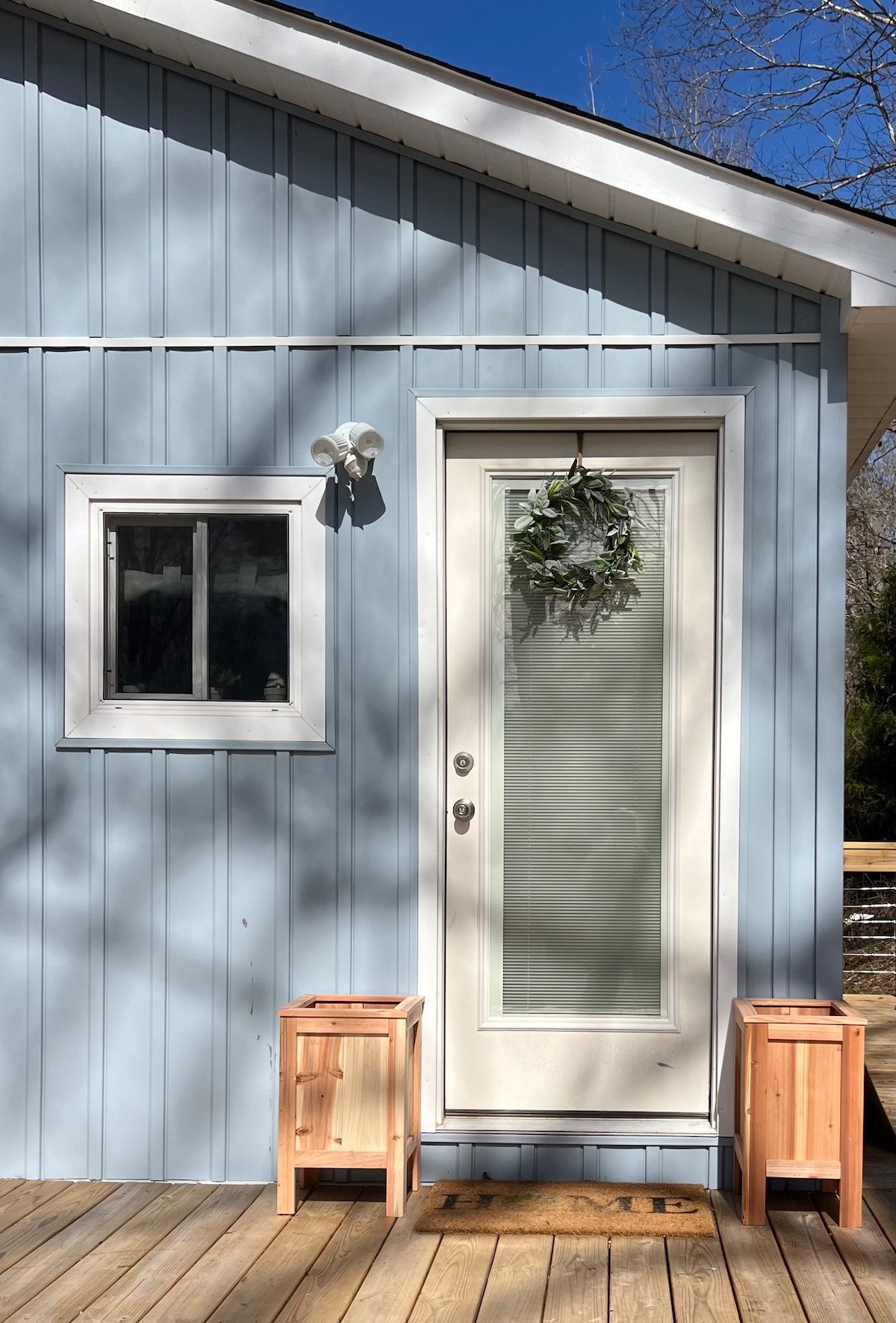 The exterior of a charming blue home is displayed, featuring a welcoming door framed by a simple wreath. A small window is positioned to the left, while two wooden stools sit on the deck in front of the entrance, enhancing accessibility.